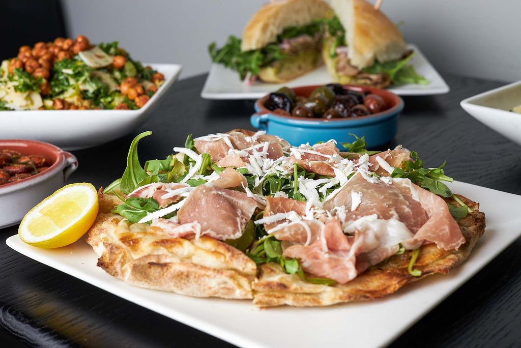 Photo of a prosciutto, arugula and cheese pizza on a white plate. Other dishes are seen on the square black table, including a small blue bowl of olives, a sandwich and a salad with roasted chickpeas on top.