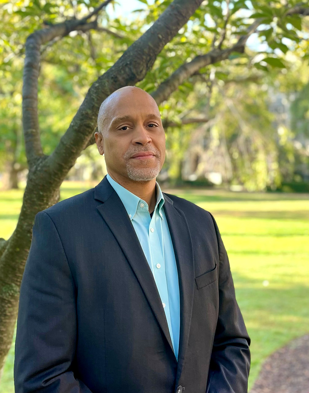 Photo of Scott Taylor. A bald black man with a neatly trimmed silver beard and mustache poses and looks directly at the camera. He wears a navy blazer and teal collared shirt and stands in front of a tree in a green field.