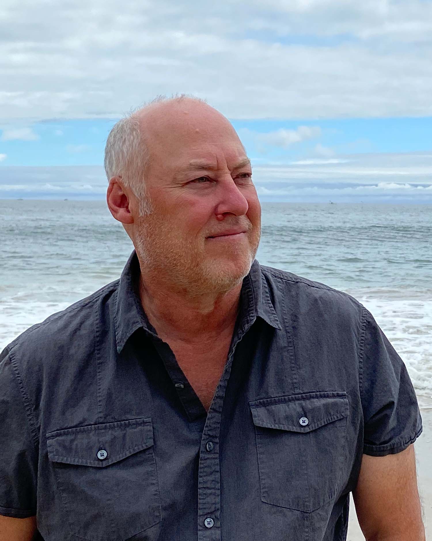 Photo of Stewart O’Nan, a middle age white man, who wears a dark gray short-sleeve button-down shirt and stands at the shore of a beach. He looks off to his left, and has short white hair and facial hair. The sky behind him is light blue with gray clouds.