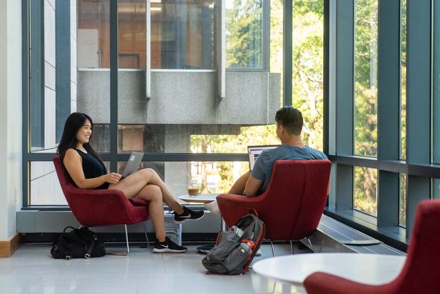 Photo of Melissa Lee (Questrom’23), left, and Rex Wang (Questrom’23) taking on their summer remote internships in the comfort of LAW’s McCausland Commons August 4. Both are young AAPI people and sit in red chairs with their laptops balanced on their laps. The chairs are situated at the end of a window-ed hallway looking out onto trees on BU beach. Their backpacks are seen on the ground.
