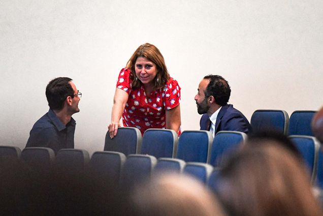 Photo of Mary Skipper, Boston Public Schools' incoming superintendent talks to Mike Sabin, (left), Boston Public School's director of school transformation and Efrain Toledano, region 6 school superintendent. Skipper is a White woman, with short blondeish hair and dangling earrings; she wears a red shirt with white flowers. She leans over to speak to Sabin and Toledano who wear blue suit jackets and blue button-downs and sit in blue lecture hall seats.