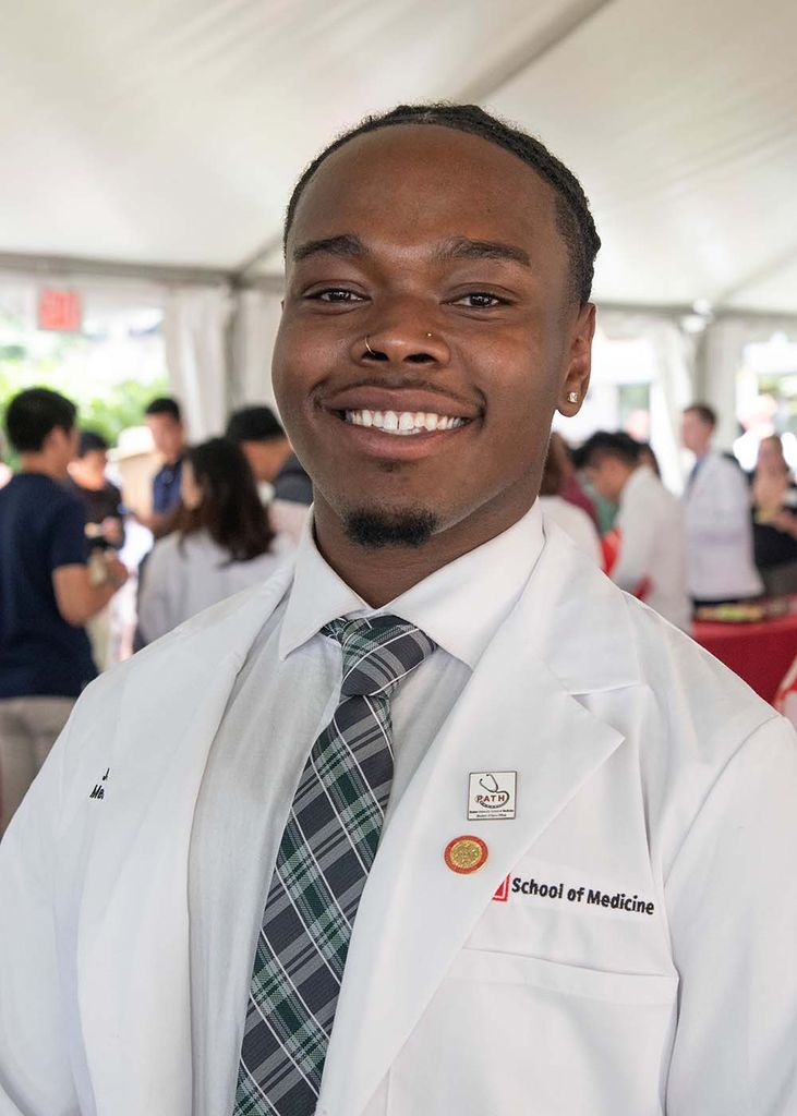 Photo of Justin Grant (MED’26), a young Black man with nose piercings who smiles and wears his white coat and a tie. He has a goatee and mustache and his hair is either short or pulled back. Other attendees of the ceremony can be seen standing underneath a white tent behind him.