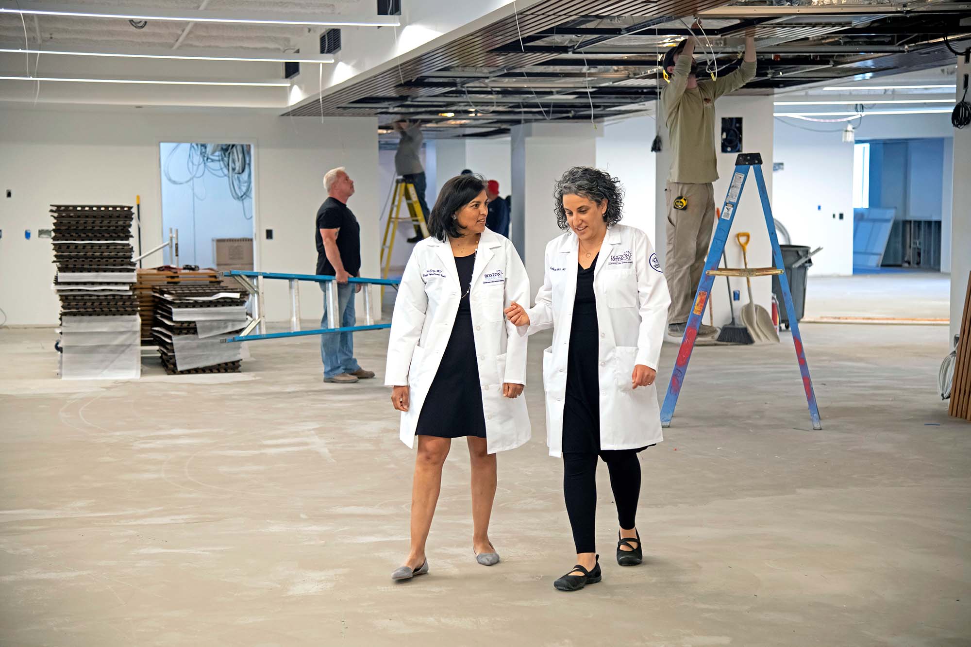 Photo of Priya Garg (left), MED associate dean for medical education, and Molly Cohen-Osher, MED assistant dean, touring the new Learning Center during construction earlier this summer. Two women wearing white lab coats chat and walk through a room under construction.