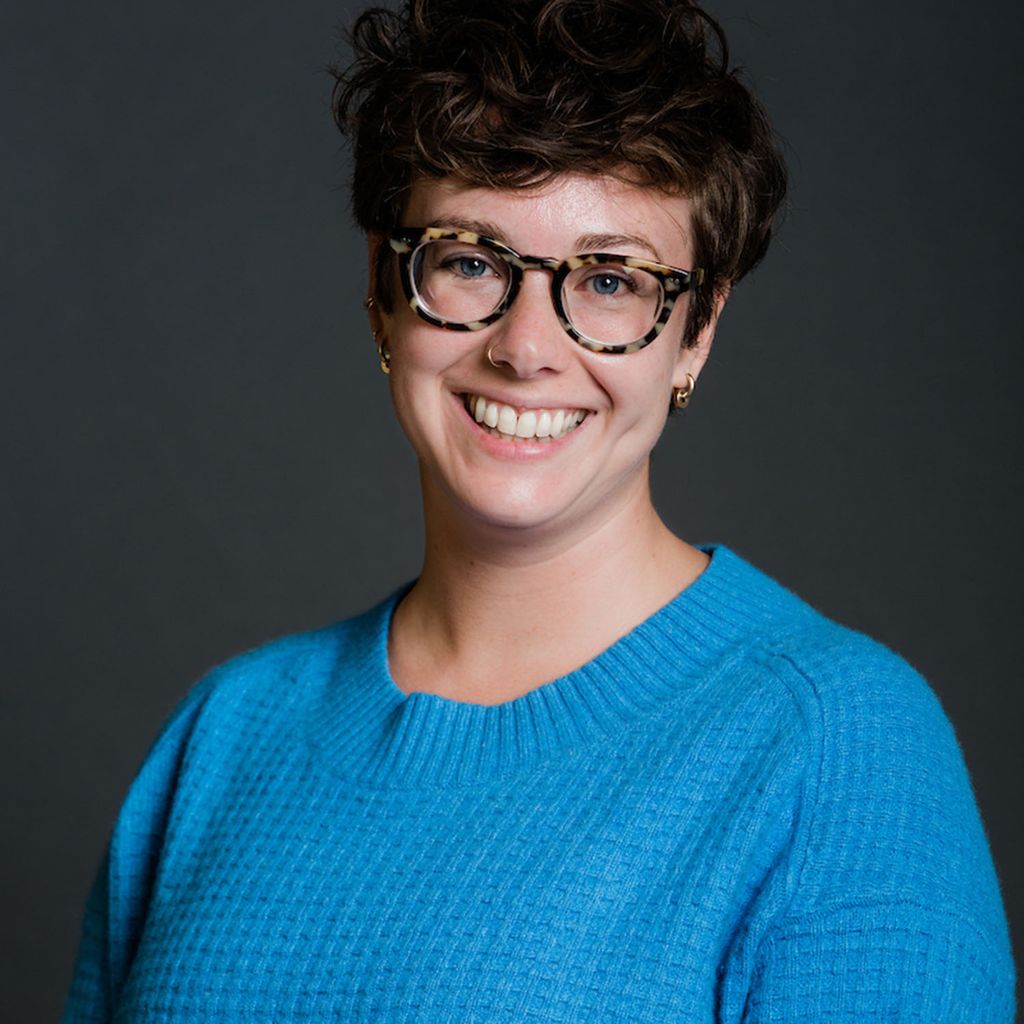 Photo: Headshot of Molly Callahan. A white woman with short, curly brown hair, wearing glasses and a blue sweater, smiles and poses in front of a dark grey backdrop.