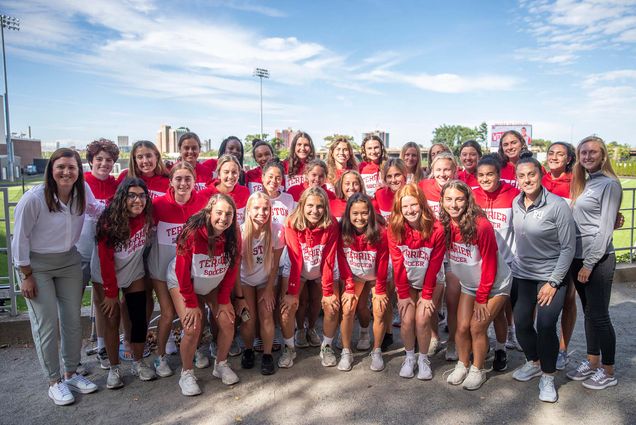 Group photo of the 2022-2023 Women's Soccer team. On the far left stands new coach Casey Brown. She wears a white collared shirt and grey pants as the teams wears red, white, and grey "Terrier Soccer" sweatshirts and grey shirts. Two athletic trainers stand tot he far right wearing grey half-zips and black pants. Entire group is smiling and posing for photo.