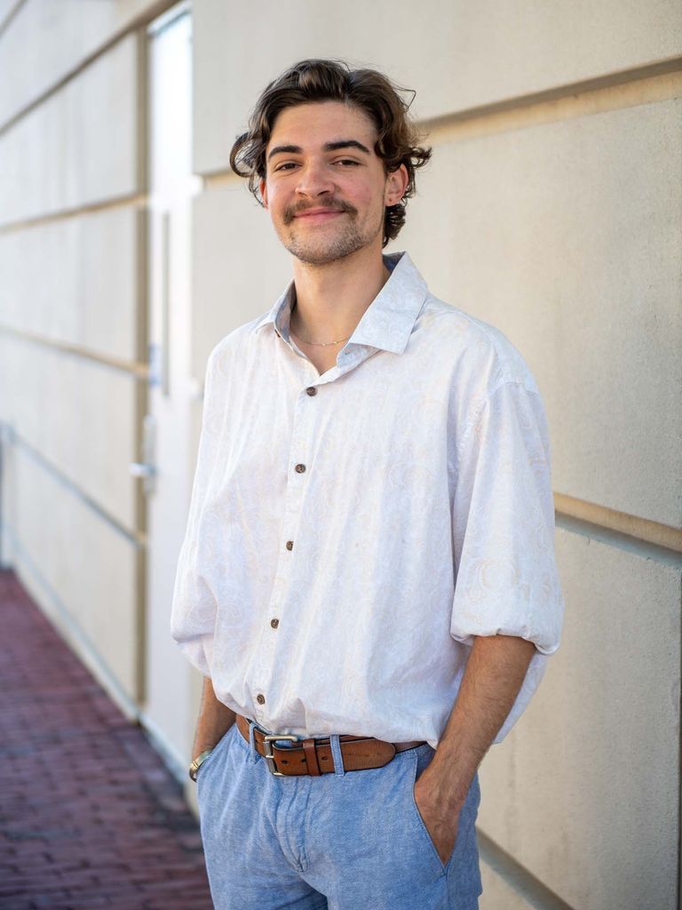 Photo of Hofmann, a young White man, who has short wavy brown hair and a mustache. He wears a short-sleeve white button down and light blue pants with a brown belt. He smiles and stands outside one of the sandstone walls outside of city hall.