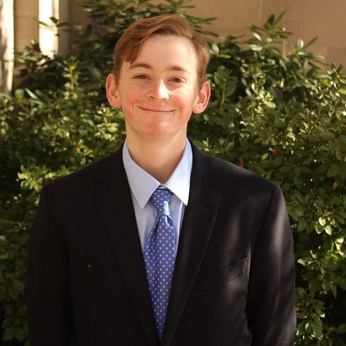 Photo: Headshot of a young white man wearing a black suit, blue tie, and light blue collared shirt. He smiles with hands clasped behind his back in front of greenery.