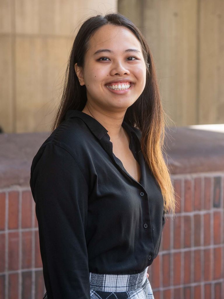 Photo of Situ, a woman of color with long brown and blonde ombre hair who wears a black long sleeve shirt and a black and white plaid shirt. She stands outside city hall in front of a brick half wall and smiles wide.