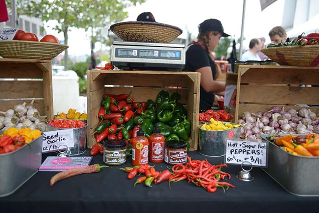 Photo of a vendor's table a the local food festival. A wooden box filed with hot red peppers and green bell peppers is seen at center; a box of garlic is seen at right. There are a few bottles of hot sauce in front of the peppers. Vendors and customers are seen blurred in the background.