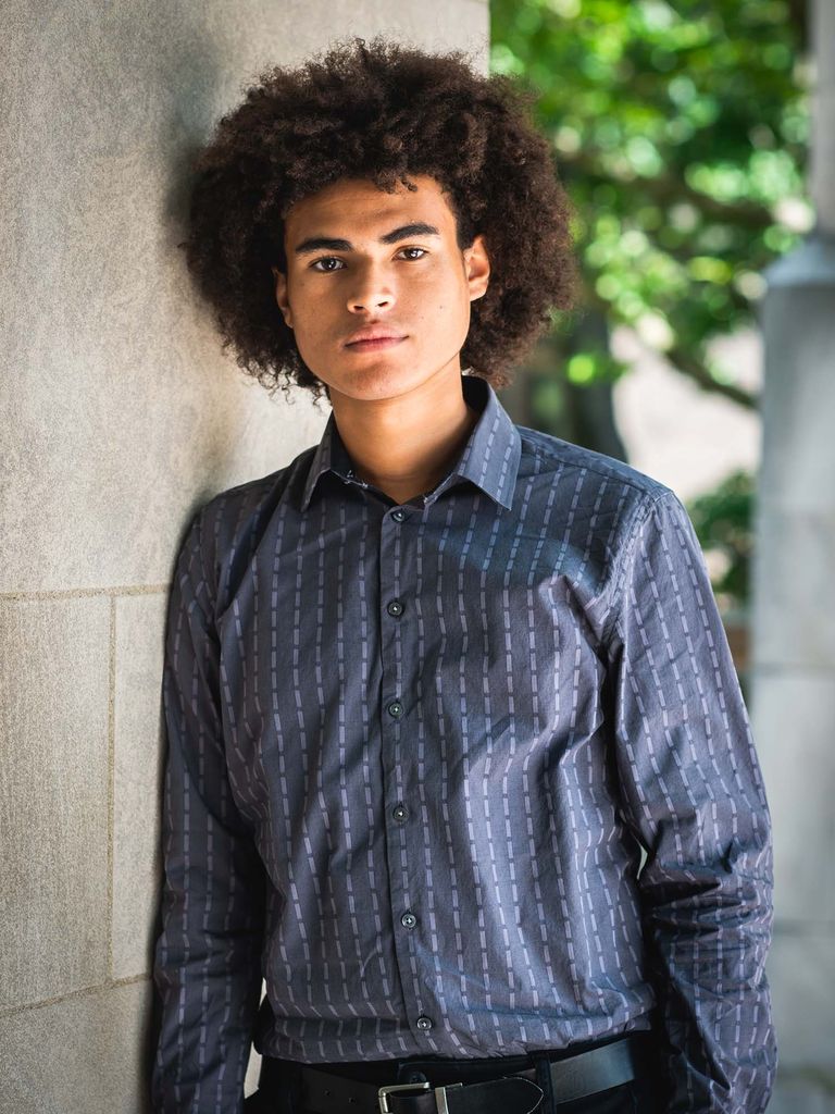 Photo of Daffin, a young man of color with long, tightly curly hair, wears a bluish gray button down and black pants. He leans against a wall and makes a serious/determined face; greenery is seen blurred behind him.