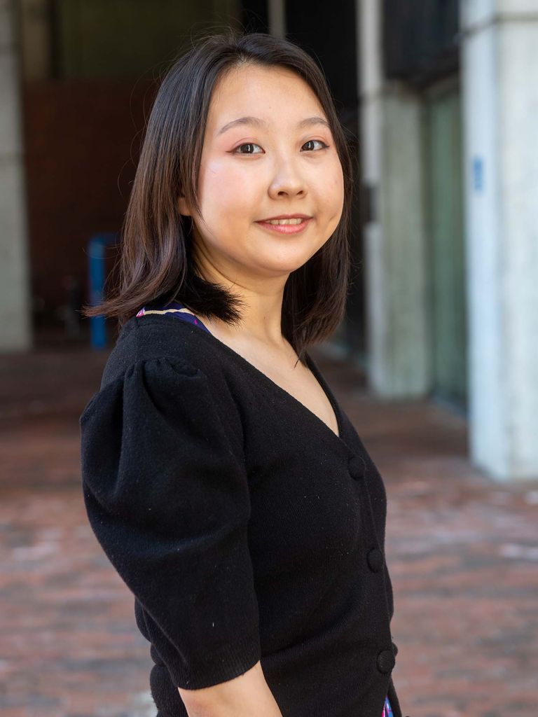 Photo of Wang, an AAPI woman with shoulder-length black hair who wears a black v-neck sweater and a floral skirt. She half smiles and is seen standing outside on the brick sidewalk in front of city hall, which is seen blurred in the background.