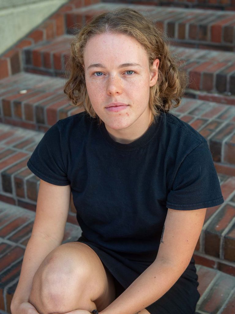 Photo of Dzialas, a White woman, wears a knee-length black dress and has short, wavy strawberry blonde hair. She sits on the brick steps outside of city hall and holds her knee in one hand. She has a serious and/or determined look on her face.