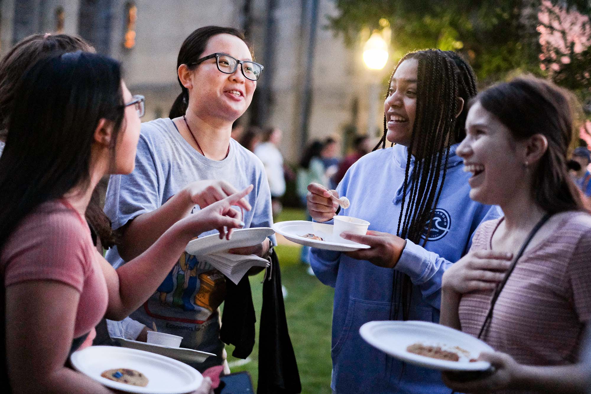 Photo: Jessica Lam (COM'24), (left), Queena Lau (CAS'24), Cat Keane (CAS'25), and Shumita Littlefield (CAS'24) talk and laugh while enjoying their dinner at BU Beach food fest. Two Asian women, a black woman with long braids, and two white women laugh as they hold plates of food and eat during dusk.