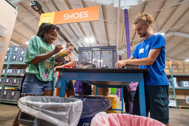 Photo of FYSOP participants Karrington Riley, (CAS’26), left, and Silvana Spagnuolo (COM’26) cleaning up donated shoes before sorting them by size at Cradles to Crayons in Newton. A young black woman on the left smiles as she cleans shoes across from a young white man (right) doing the same.