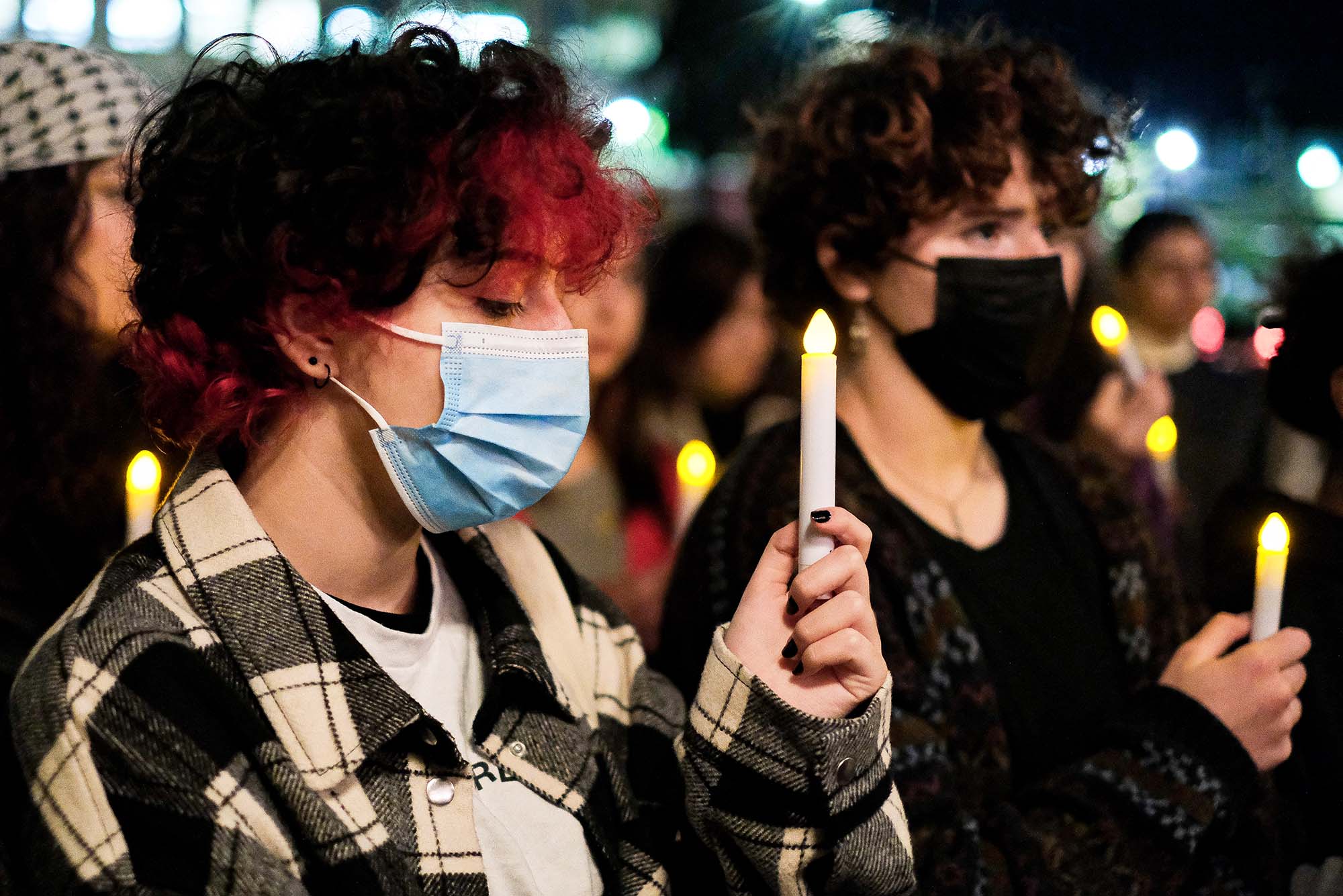 Photo: A student holds her candle during the candlelight vigil event on Marsh Plaza Sep 29th. A masked student stands with eyes closed and electric candle raised as she stands next to another masked individual to her right. A blurry crowd of other participants holding candles can be seen behind them.