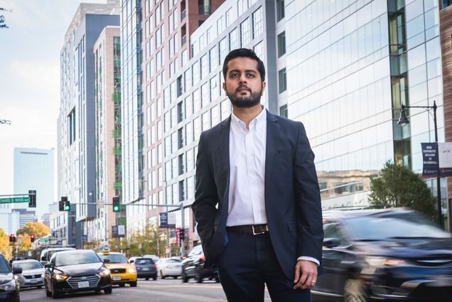 Photo: SHA faculty member Makarand Mody poses for a photo on Boylston Street in Boston. Modt poses in a suit ensemble in front of a city scene of high rise buildings and busy streets.