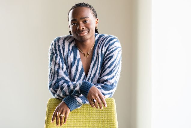 Photo of Chike Asuzu posing in a white room next to a large window. A Black person wearing a blue and white leopard-striped long-sleeved sweater leans forward on a yellow chair placed in front of them.