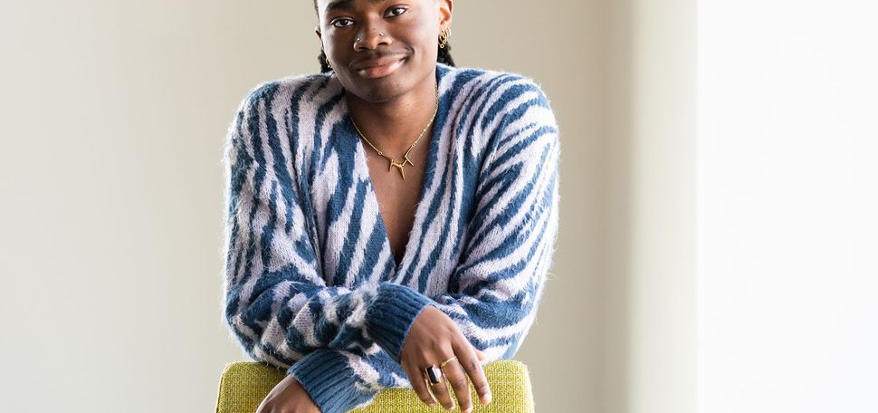 Photo of Chike Asuzu posing in a white room next to a large window. A Black person wearing a blue and white leopard-striped long-sleeved sweater leans forward on a yellow chair placed in front of them.