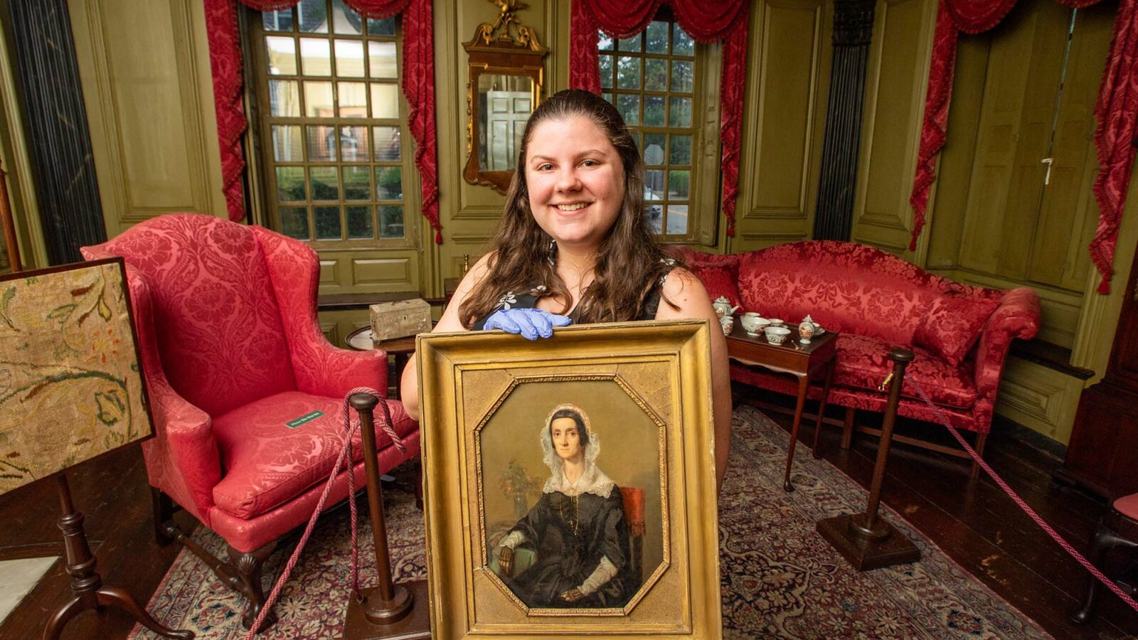 Photo: American Studies PhD student MaryKate Smolenski stands and smiles in the northeast parlor and holds a portrait of Mary Robinson Hunter at Hunter House in Newport, RI. A young white woman stands in an ornate gold and red decorated room holding an old portrait in a gold frame with gloved hands.