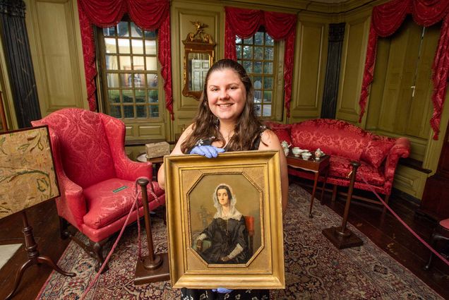 Photo: American Studies PhD student MaryKate Smolenski stands and smiles in the northeast parlor and holds a portrait of Mary Robinson Hunter at Hunter House in Newport, RI. A young white woman stands in an ornate gold and red decorated room holding an old portrait in a gold frame with gloved hands.