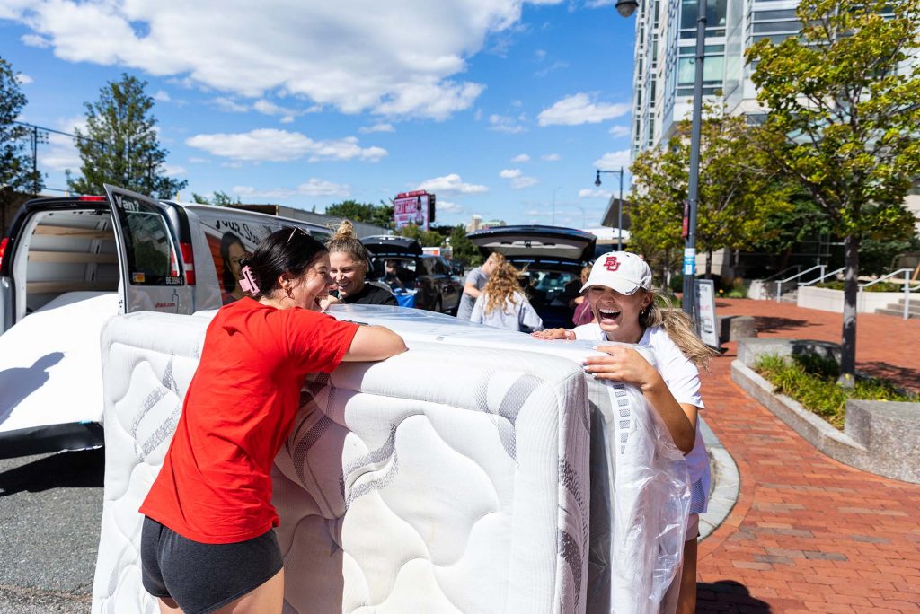 Photo: Students move into StuViII. A young Asian woman and a young white woman laugh as they slowly move a large white mattress from a van.