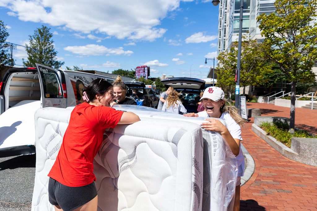 Photo: Students move into StuViII. A young Asian woman and a young white woman laugh as they slowly move a large white mattress from a van.