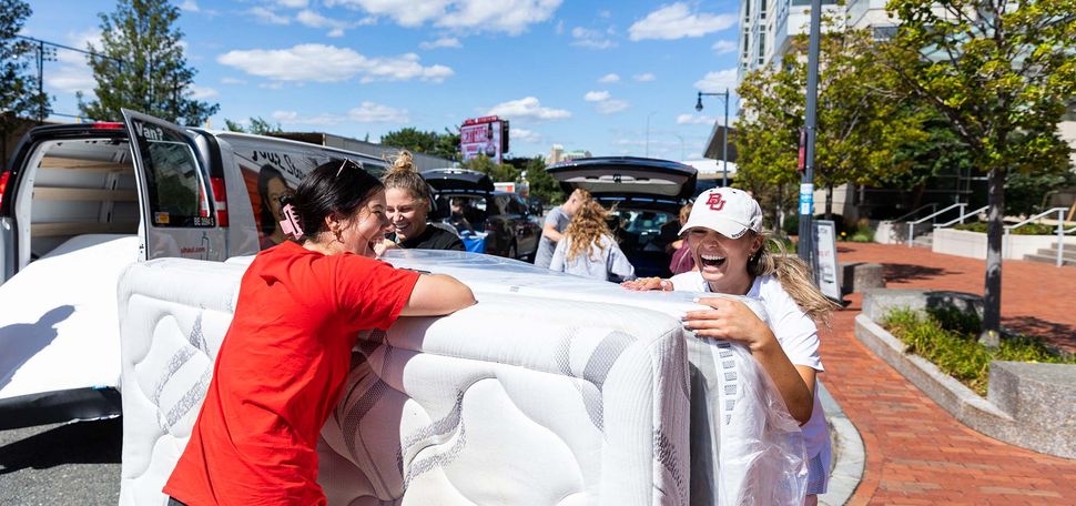 Photo: Students move into StuViII. A young Asian woman and a young white woman laugh as they slowly move a large white mattress from a van.