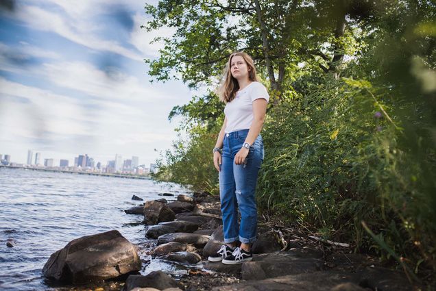 Photo: Natalia Sawicka, a young white woman with long blonde hair, poses for a photo on the bank of the Charles River. The city of Boston can be seen distantly in the background.