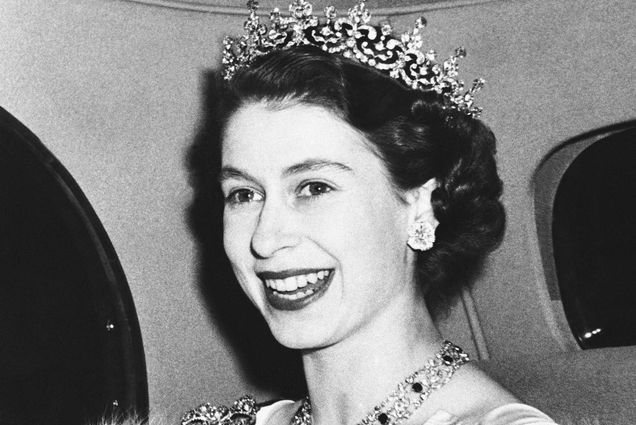 Black and white photo of Princess Elizabeth smiling broadly as she arrives at the French Embassy in London's Kensington Palace Gardens. A young white woman with coifed dark hair and wearing a jeweled tiara smiles broadly and looks to the left of the camera.