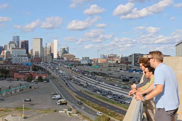 Photo: BU School of Public Health researchers (from left) Christopher Rick, Jonathan Buonocore, and Chad Milando are hoping to show how a functioning public transport system is an essential public health resource. Three men stand at a railing and look off into the distance overseeing a busy, congested highway