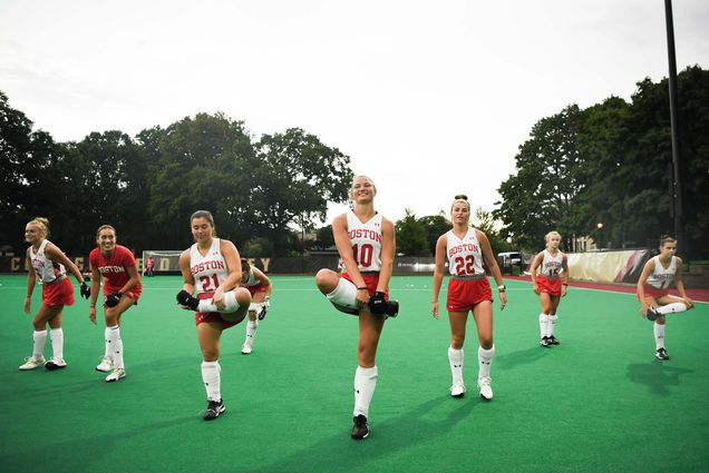 Photo: A group of BU women's field hockey players stand next to each other as they hold one leg up and hold it in a figure four. They all wear red shorts and white tank tops and stand on a green turf field.