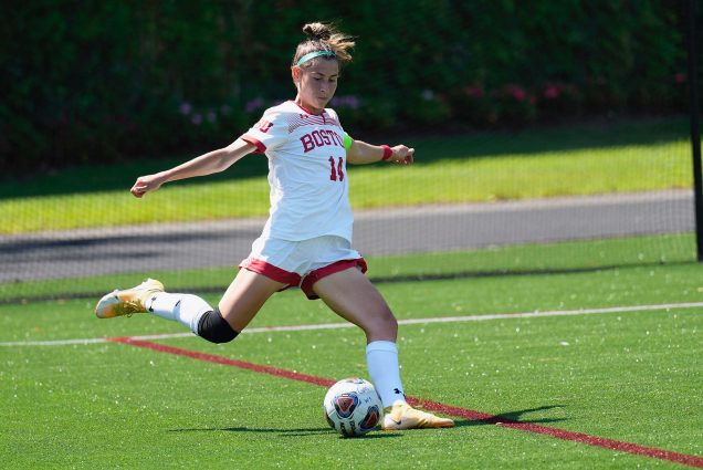 Photo of 2022 Women’s soccer captain Amy Thompson. A young white woman wearing a White BU soccer uniform is shown mid-kick to a soccer ball on the soccer field.