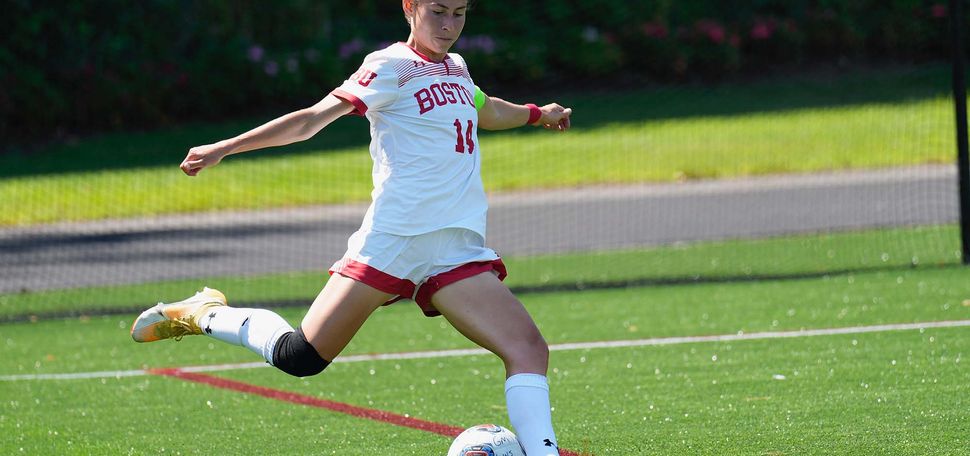Photo of 2022 Women’s soccer captain Amy Thompson. A young white woman wearing a White BU soccer uniform is shown mid-kick to a soccer ball on the soccer field.