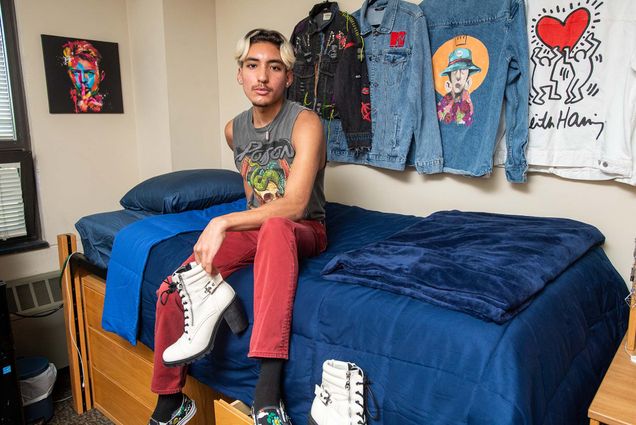 9/2/22 -- Boston, MA Matthew Garcia (Questrom’26) in his Claflin Hall dorm room Photo by Cydney Scott. Matt sits on a twin xl bed on risers with a blue bedspread. Holding a pair of white heels, Matt is also wearing red pants and a tank top with a fashionable graphic. Collared shirts of various designs hang on the wall behind.