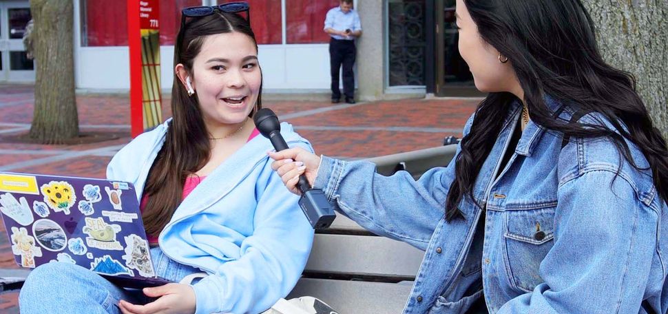 Photo of Irene Kim interviewing a student in front of the BU student union. Two female students sit on a bench facing each other. The student on the right holds a microphone up as the student on the left speaks into it.