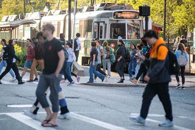 Image of a group of students crossing the large intersection across from the Howard Thurman Center. Students walk in both directions. A trolley car from the T's is seen behind the crowd. Green line.
