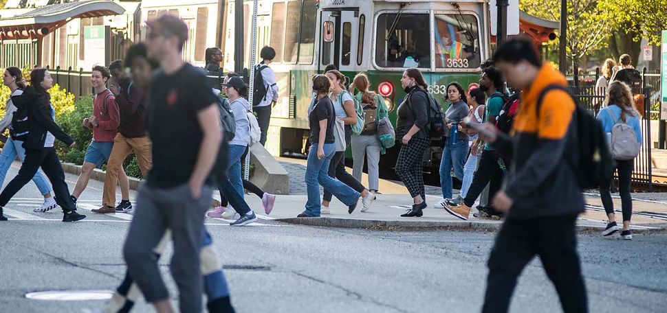 Image of a group of students crossing the large intersection across from the Howard Thurman Center. Students walk in both directions. A trolley car from the T's is seen behind the crowd. Green line.