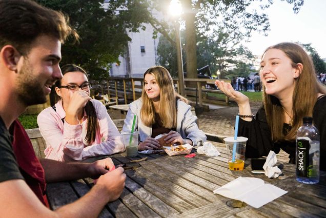 Photo of Mank Rutu (ENG'24), (left) a White man with brown hair, Nicole Zaju (ENG'24), a lightskinned woman with glasses and long brown hair, Niana Geoffrion (Questrom'24), a White woman with long blonde hair and a gray sweatshirt, and Marianne Palmieri (ENG'24), a White woman with long brown hair, chatting while having their dinner on BU Beach. They sit at a wooden table and most wear long-sleeves. Boba is seen in plastic cups on the table and the group smiles and laughs. It looks to be dusk outside.