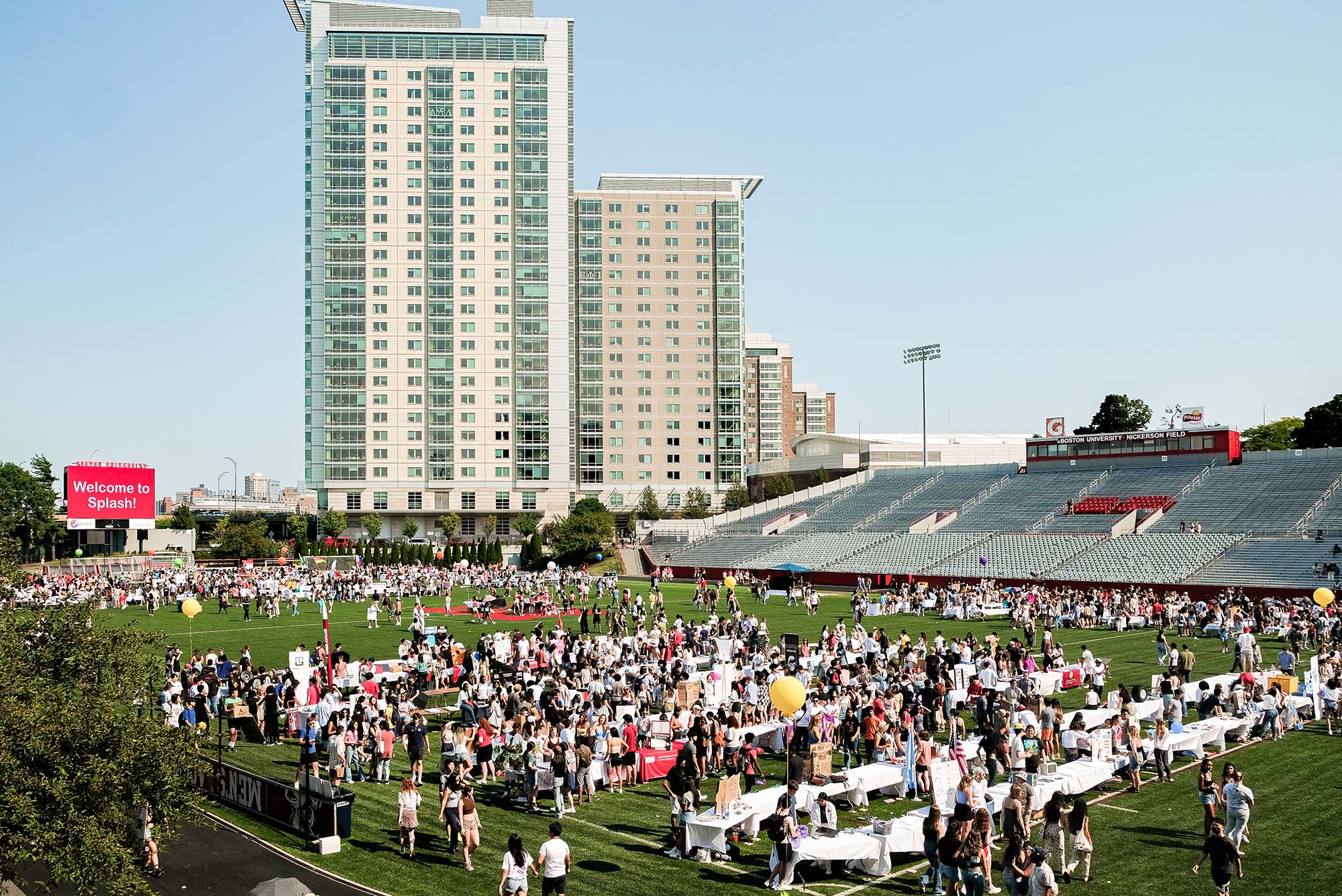 Wide photo of Nickerson field during the annual SPLASH event. Students walk across the field to look at the tables set up by BU student clubs. STUVII is seen in the background; the sky is blue and clear.