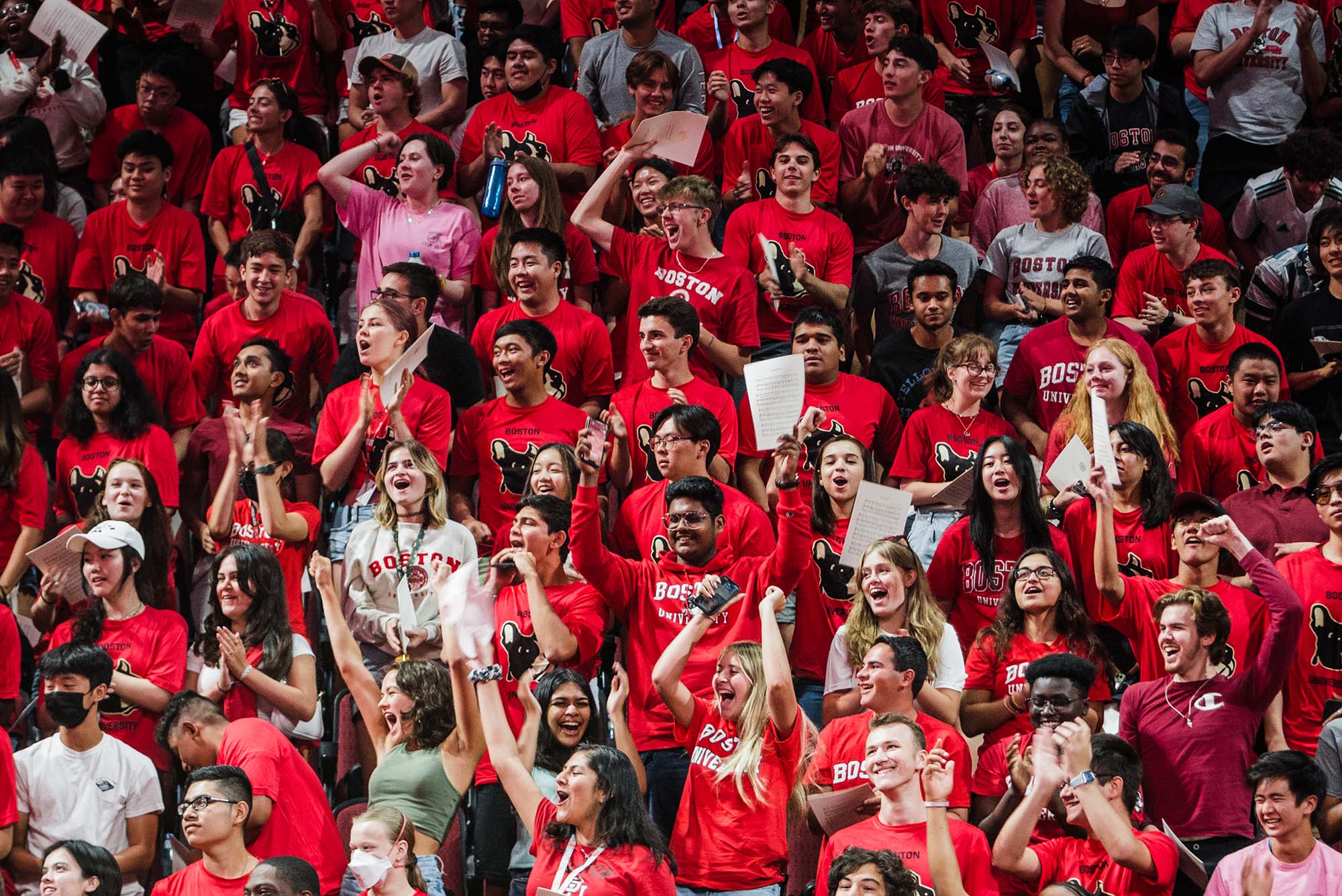 Photo of a diverse group of students cheering during the matriculation event in Agganis Arena. Almost all of them wear red BU tshirts, with either the words "Boston University" or an image of a terrier on them. Many also hold papers in their hands.
