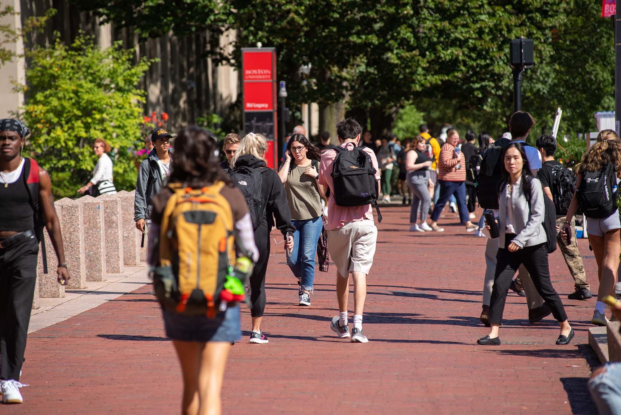 Photo of students walking up and down Comm Ave's brick sidewalk by Marsh Chapel. Many wear backpacks and it is a sunny day.