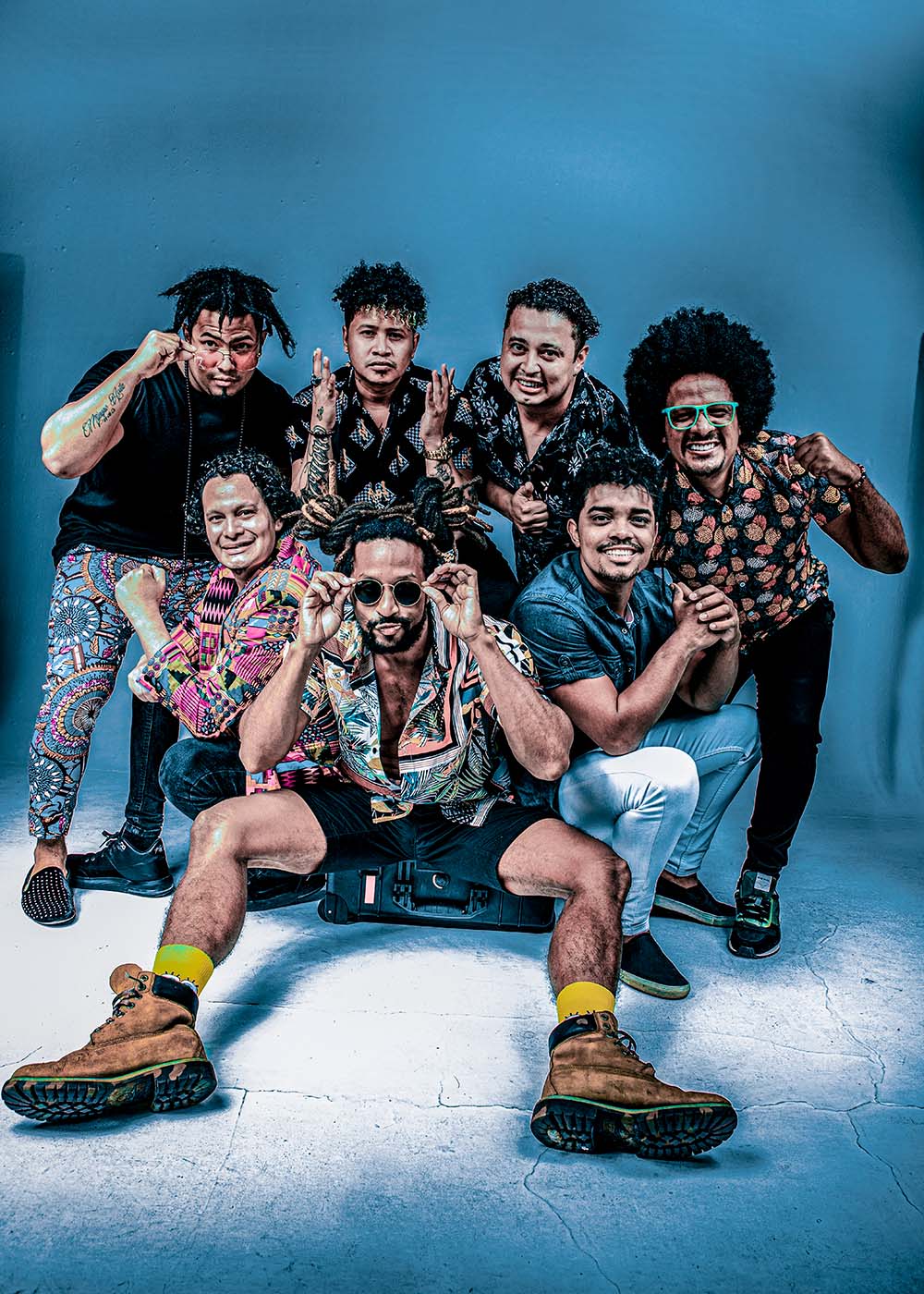 Photo of champeta band The Bazurto All-Stars of Bogota, Colombia. The band consists of seven people of color who are dressed in bright patterns and wear natural hair styles. Four crouch in a huddle and three sit on what looks like a small suitcase in front. The background is cement lit blue.