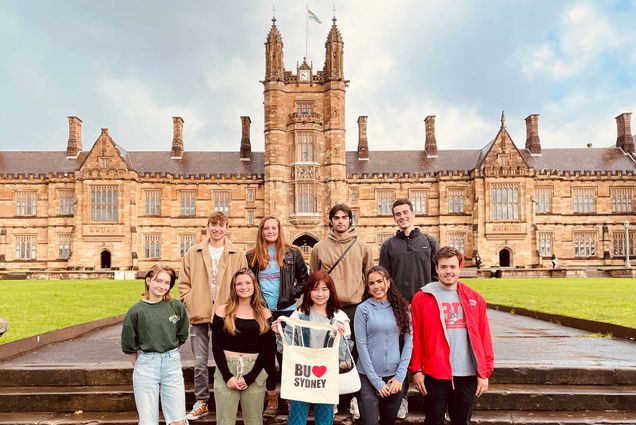 Photo of a group of nine BU students posing in front of a European style palace building with a central tower. An AAPI student at center holds a tote that reads "BU :heart: Sydney." They wear sweatshirts and pants and the day looks slightly overcast.