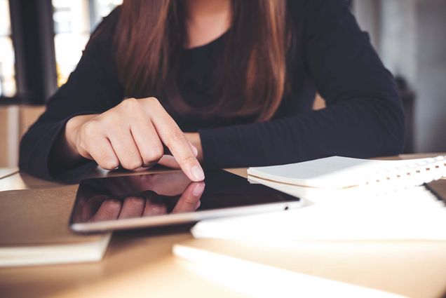 Photo of a person with shoulder length brown hair using their right hand to point at a black tablet screen. The tablet is next to a notebook on wooden table. The person wears a dark bluish sweater.