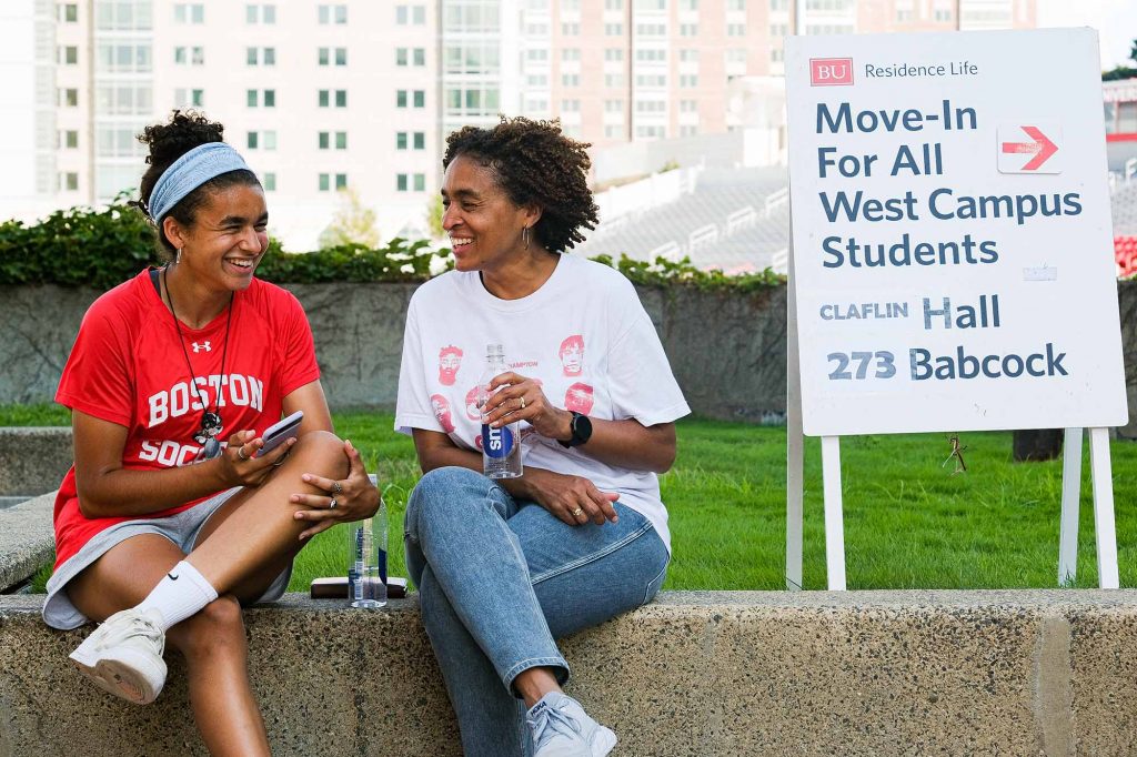 Photo: Sitting under the shade next to Rich Hall, Shayla Brown (Sargent'26), (left) takes a move-in break with her mom. Two brown women are turned towards each other as they sit on a cement bench and laugh. The sign tot he right of them reads "Move in for all West Campus Students"