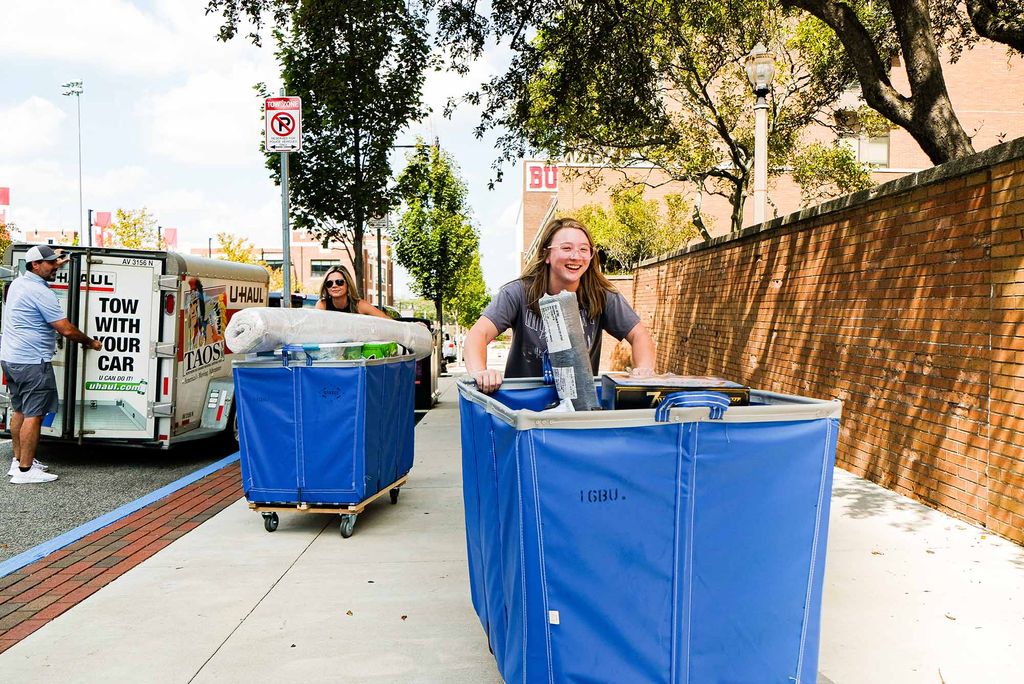 Photo: Palmer Wencley (COM'26) moves her stuff across West Campus with her parents helping in the back. A smiling, young white woman and her mother both push large blue bins filled with stuff up the sidewalk. A white father figure stands at the back of a small UHaul truck and looks on.