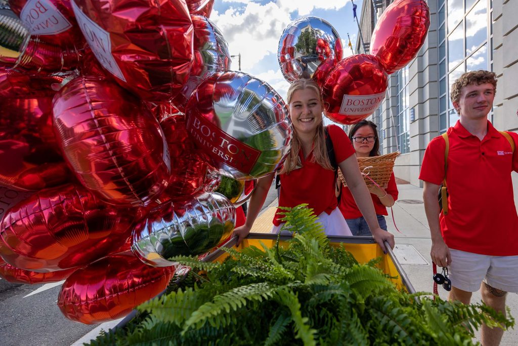 Photo: Students wearing red collared shirts  and white pants smile as they push a large yellow bin filled with fake plants down the sidewalk. They also hold a large set of red and silver Boston University helium balloons.