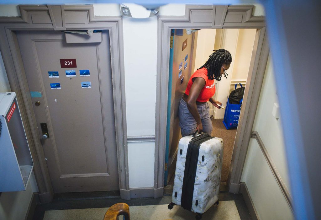 Photo: Mary Adekunle (CAS 26) moves into her dorm on South Campus. A young black woman wearing a red shirt and jeans moves a medium-sized suitcase through the door of her apartment. 