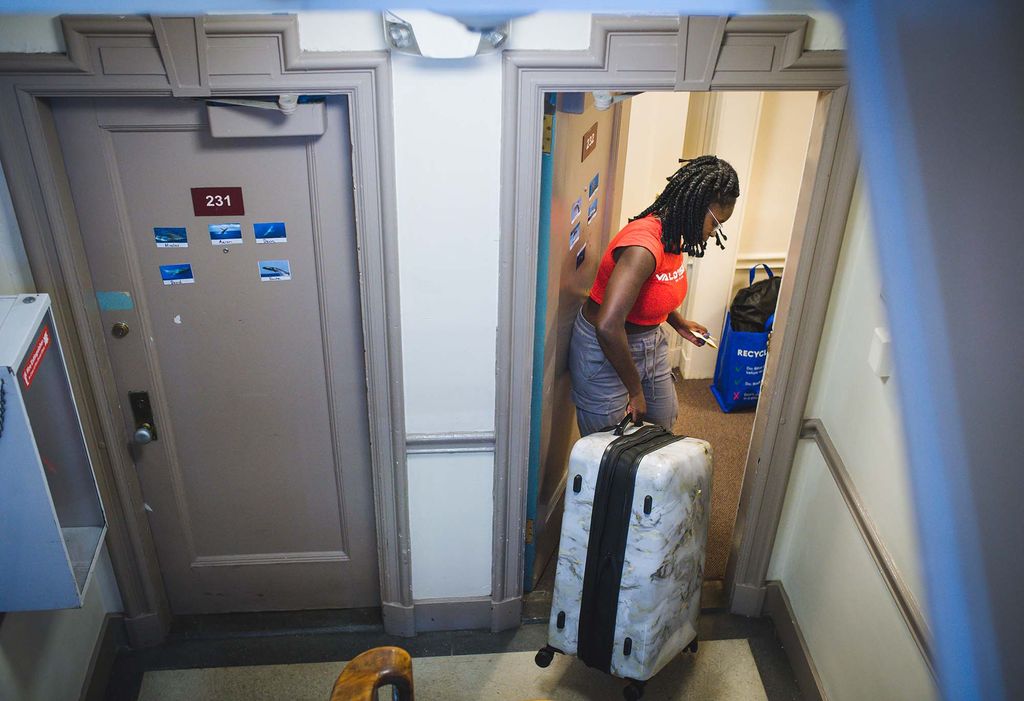 Photo: Mary Adekunle (CAS 26) moves into her dorm on South Campus. A young black woman wearing a red shirt and jeans moves a medium-sized suitcase through the door of her apartment.