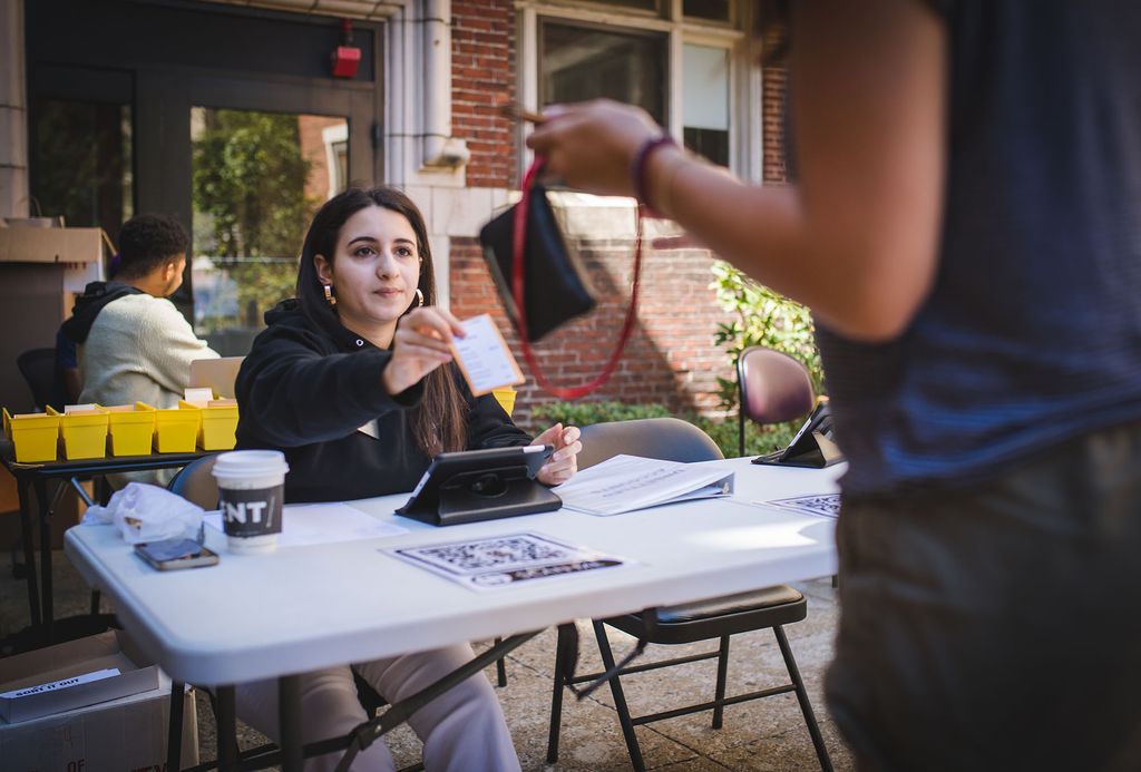 Photo: RA Natalia Kanj (CAS 22) checks in students on South Campus. A young white woman with long dark hair sits at a plastic folding table and hands keys out to incoming residents.